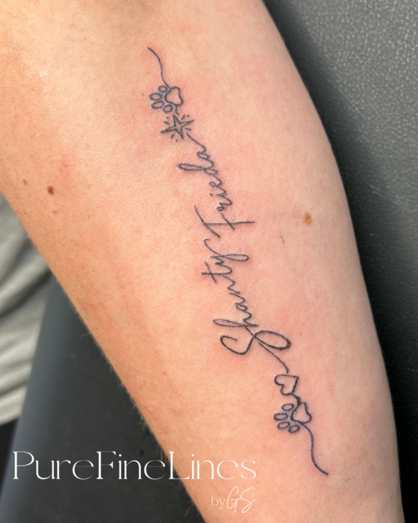 Fineline Schriftzug-Tattoo in Schreibschrift mit kleinen Pfotenabdrücken und Blüte am Unterarm, PureFineLines by GS